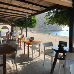 Mesas y sillas bajo un techo de paja con vista a una playa de arena y el mar. Un lugar perfecto para disfrutar de la **agua amarga donde comer**.