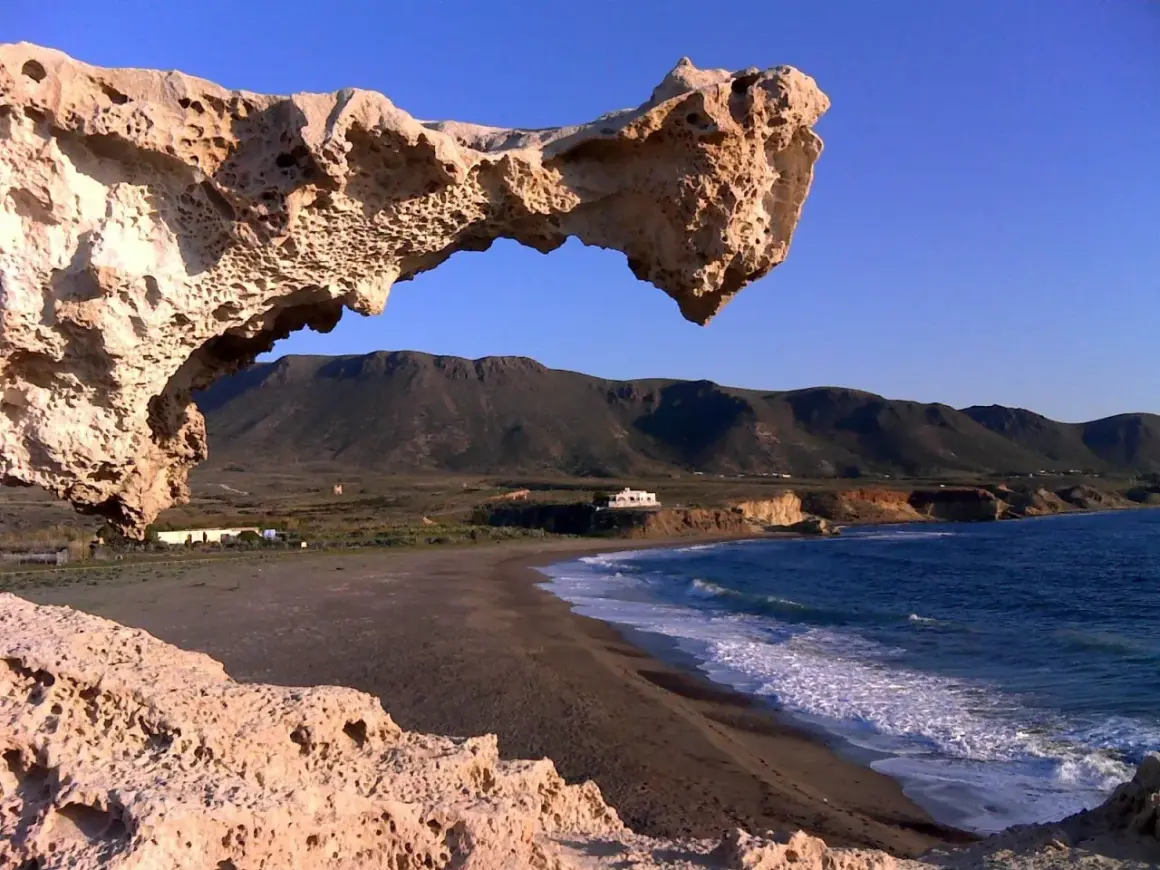 Formaciones rocosas erosionadas, como los escullos, enmarcan una playa de arena oscura y el mar azul. Montañas verdes y una casa blanca al fondo.