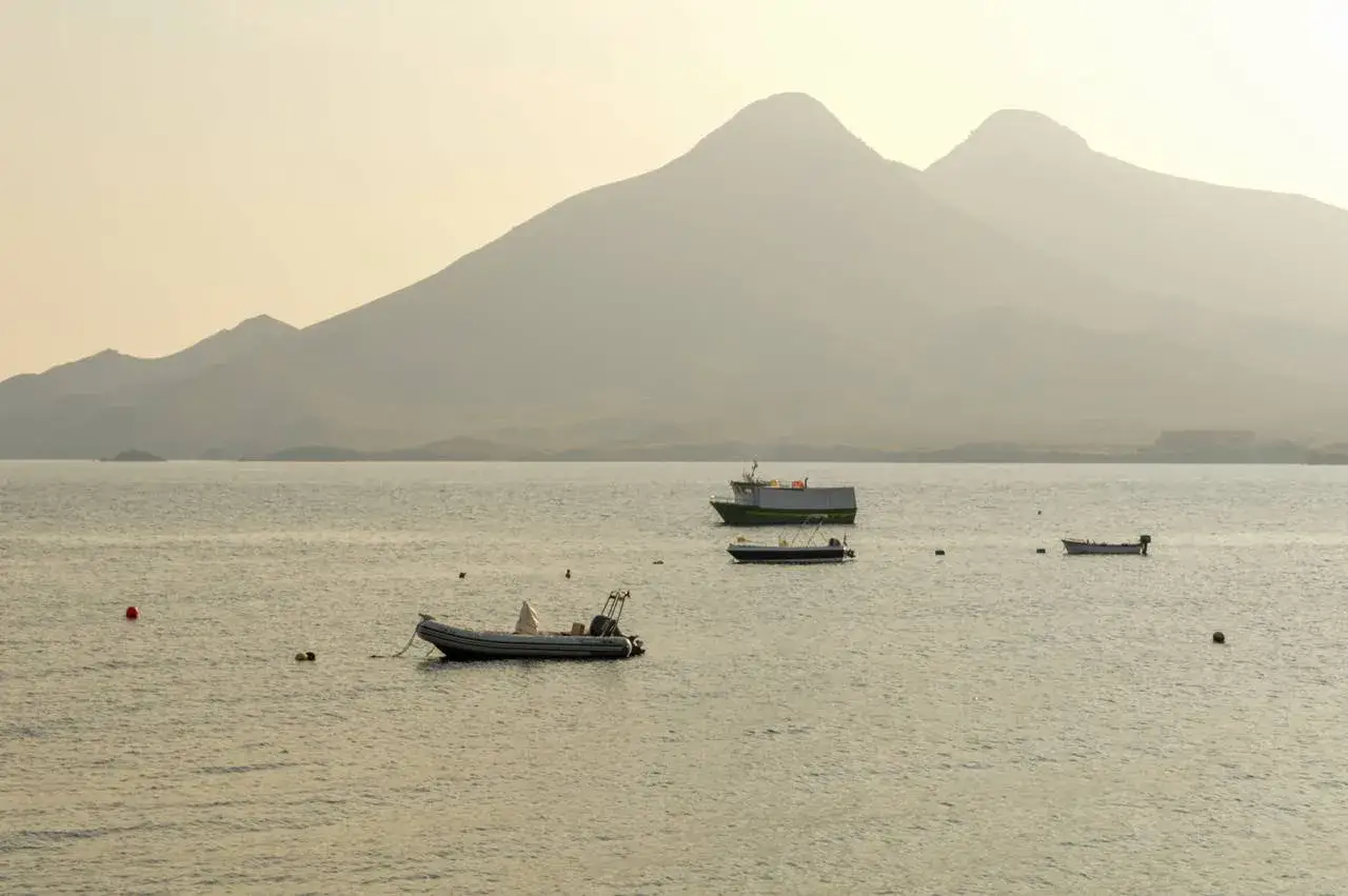 Barcos flotan en calma marina ante silueta de montañas. La isleta del Moro se vislumbra en la distancia, un refugio sereno.